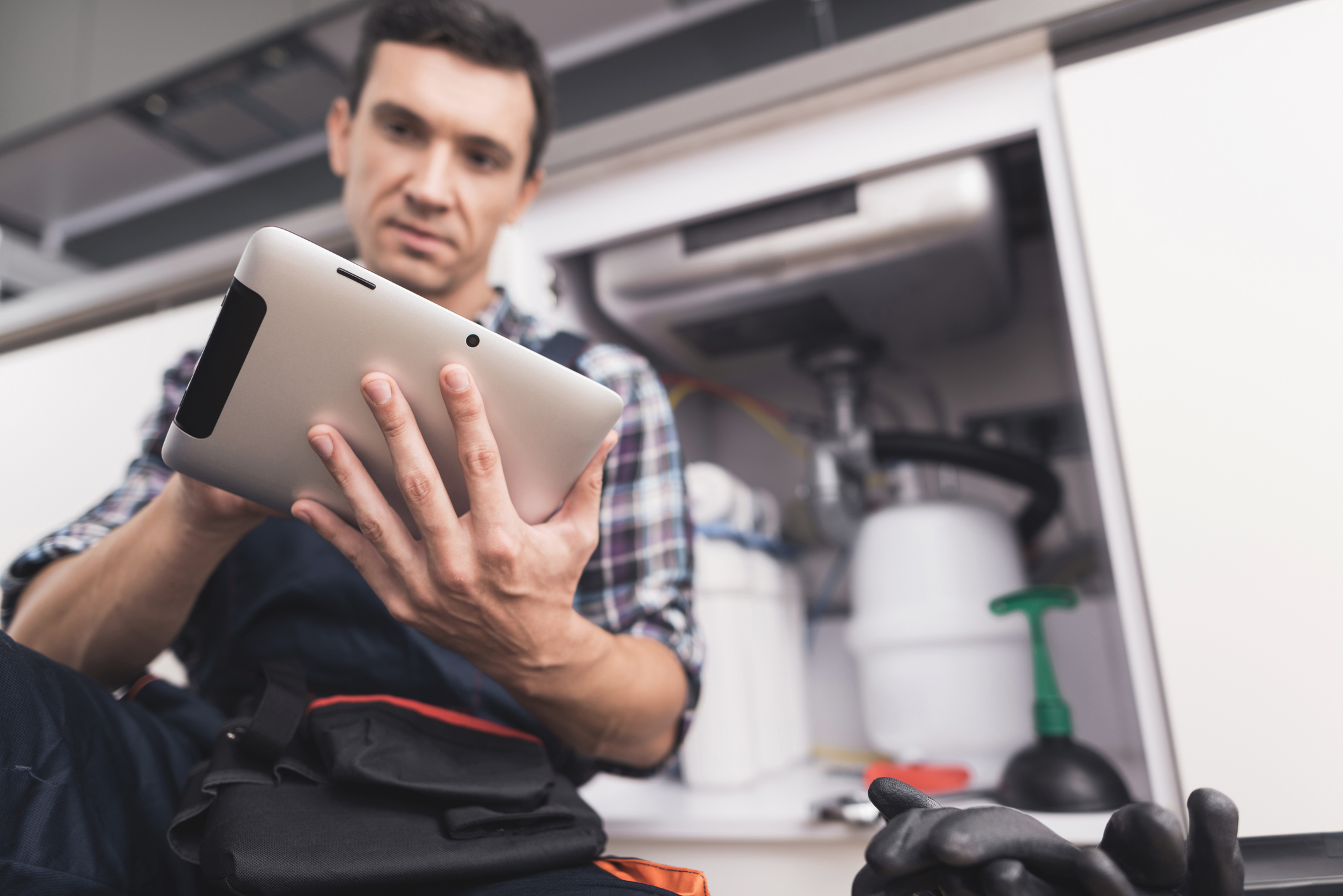 Innovative Plumbing Technologies, The plumber sits next to the kitchen sink on the floor and looks at the tablet for repair instructions.