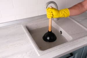 Gloved hand holding plunger against clogged sink, clearing clog, Residential Plumbing Overflows.